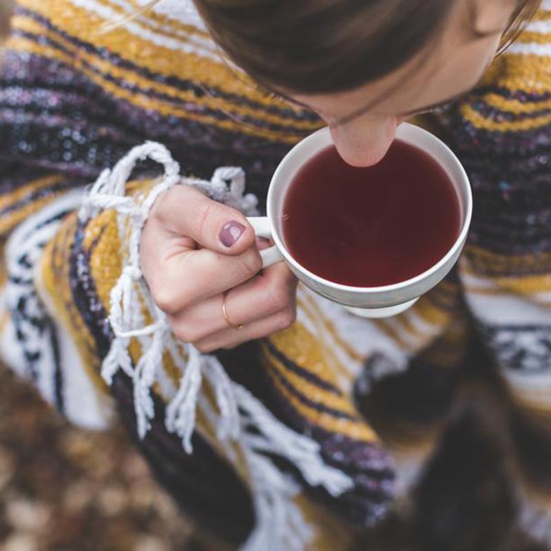 A cup of herbal tea being sipped by a dark haired lady in a poncho.