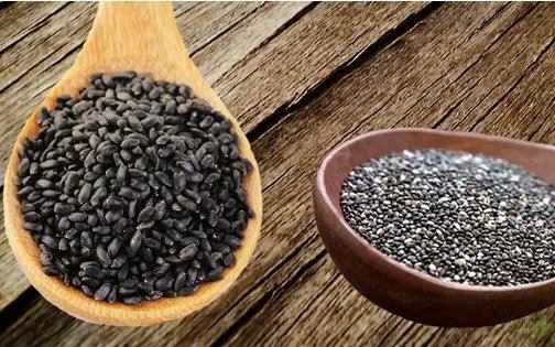 A wooden spoon of black basil seeds beside a bowl of chia seeds on a wooden board