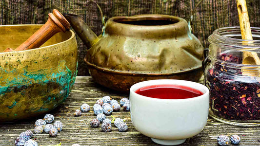 Rustic herbal tea setup with a brass mortar and pestle, vintage teapot, dried hibiscus in a glass jar, and a cup of red herbal infusion on a wooden table