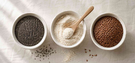 Three white ceramic bowls on a linen background: black chia seeds on the left, organic psyllium husk powder with a wooden spoon in the centre, and bronze flaxseeds on the right.