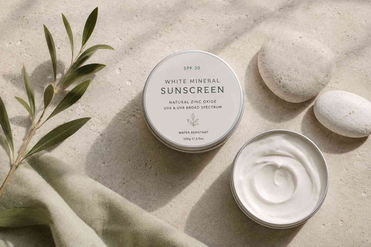 Open tin of white mineral sunscreen with natural zinc oxide, SPF 30, flat lay with smooth stone pebbles and an olive leaf sprig on a warm linen surface.
