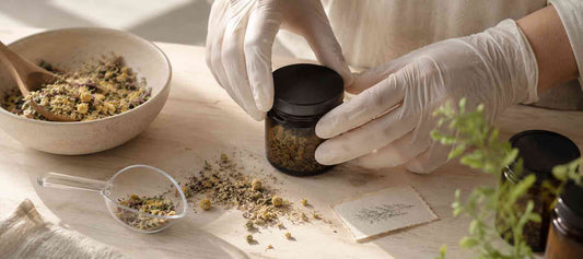 Gloved hands filling a dark glass jar with dried herbs on a pale wood surface, with a bowl of dried botanicals in the background.