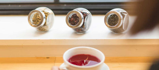 A cup of hibiscus tea on a bench with three pots of loose tea nearby, Chamomile tea, Ginger Tea and HIbiscus Tea.