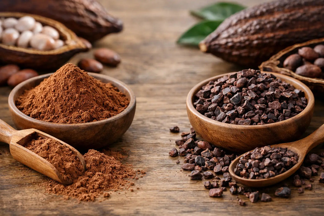 Cacao powder and cacao nibs in wooden bowls with scoops, beside whole cacao pods and beans on a wooden table.