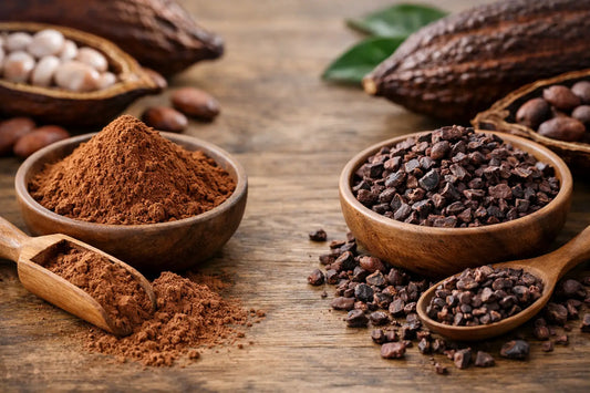 Cacao powder and cacao nibs in wooden bowls with scoops, beside whole cacao pods and beans on a wooden table.