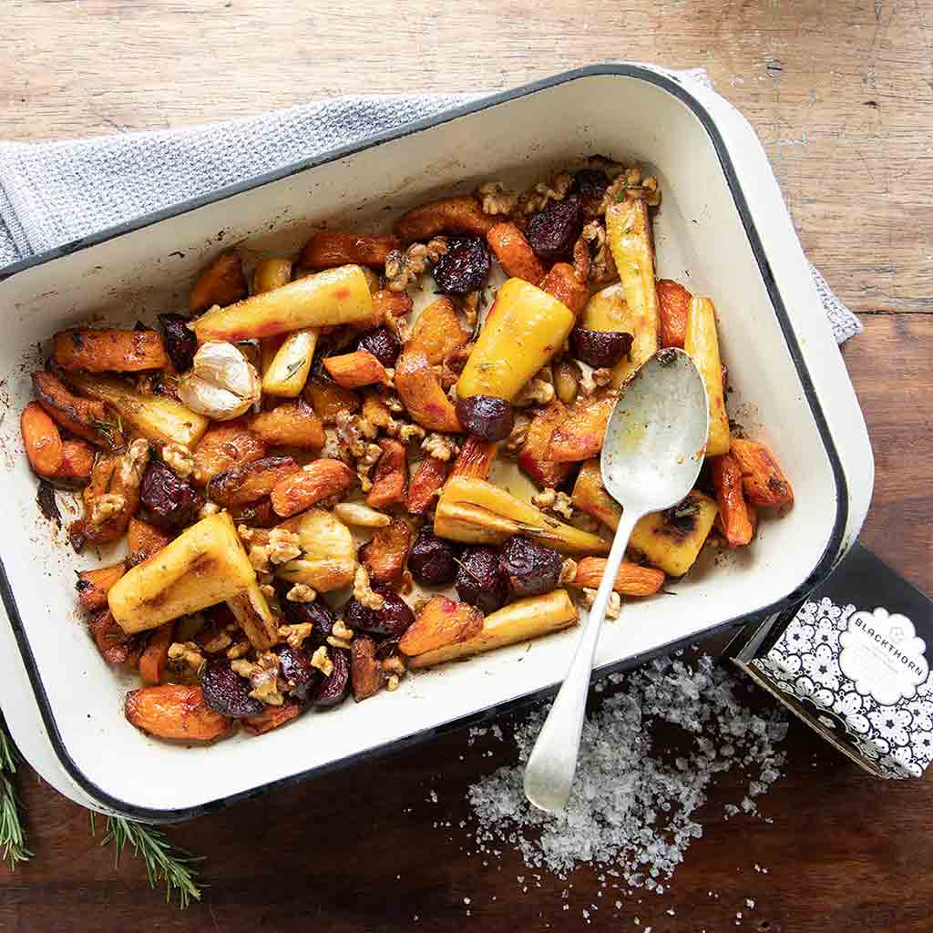 Roasted root vegetables with nuts in a white baking dish on a wooden surface with Blackthorn Sea Salt.