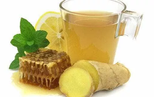 A cup of ginger tea in a clear glass cup with a fresh piece of ginger, honey comb and a mint leaf.