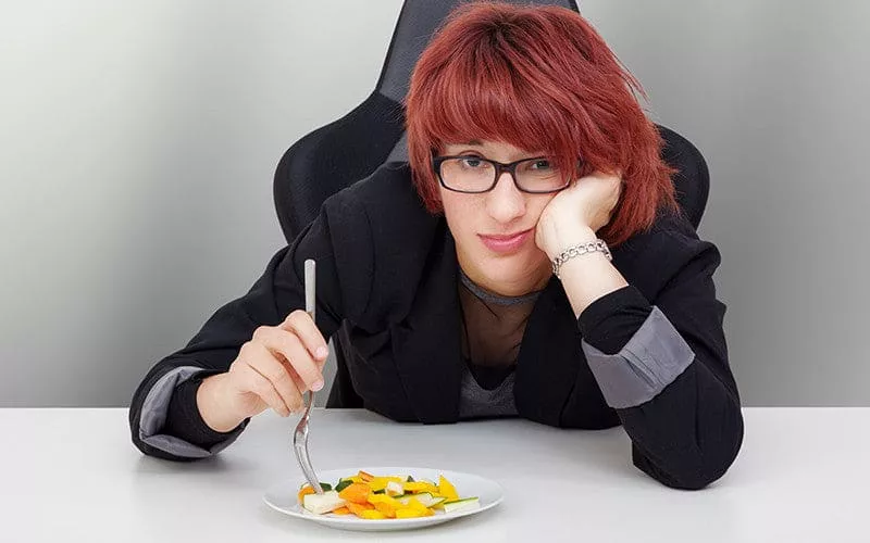 A red haired lady sat at a desk looking bored eating a plate of fresh salad.